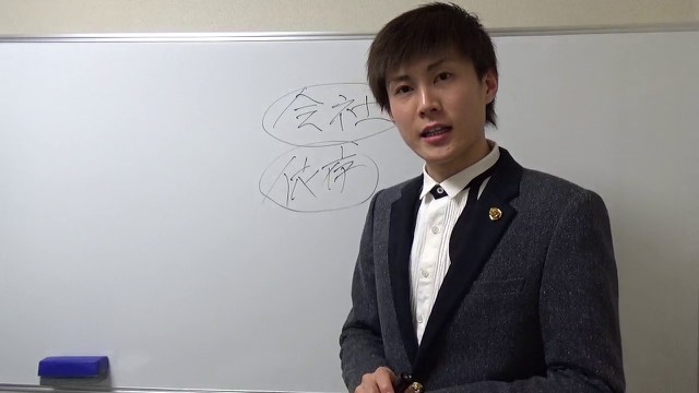 ホワイトボードに「会社」「依存」の丸囲み文字を背景に話す本人(本文の該当シーン)