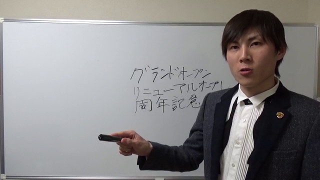 ホワイトボードの「グランドオープン」等の板書を指し示す本人