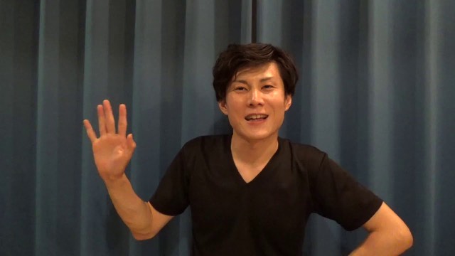 青いカーテン背景で手を挙げて話す本人(動画「トラブルを人のせいにしたら赤信号です」より)