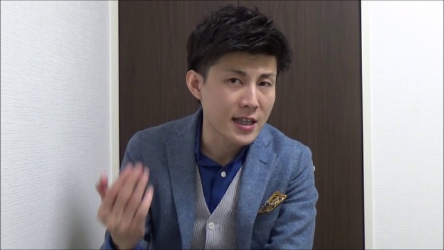 ジャケット姿で手振りしながら話す本人(動画「ネットビジネスで稼ぐコツ 環境が超重要」より)