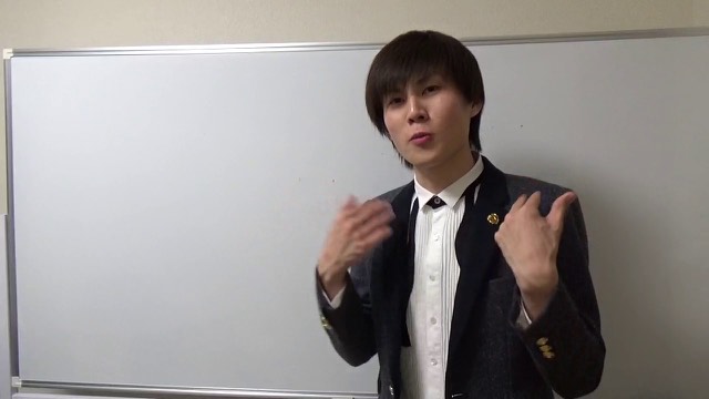 ホワイトボード前でスーツにネクタイピン姿の本人が両手を広げて話す(動画「お金持ちに会うメリット【パチスロ】【スロット】」より)