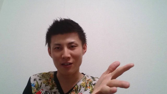 無地の壁を背景に花柄シャツ姿の本人がピースサインをして話す(動画「最近のパチスロで稼げる人になるには?【パチスロ】…」より)