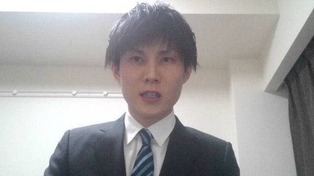 スーツにネクタイ姿で明るい室内に立つ本人(動画「弟子に応募していただきありがとうございます!」より)