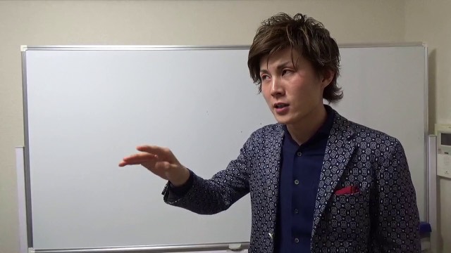 柄物ジャケットでホワイトボード前に立ち指差す本人(動画「多くのスロッターは時間が止まってる【パチスロ】…」より)