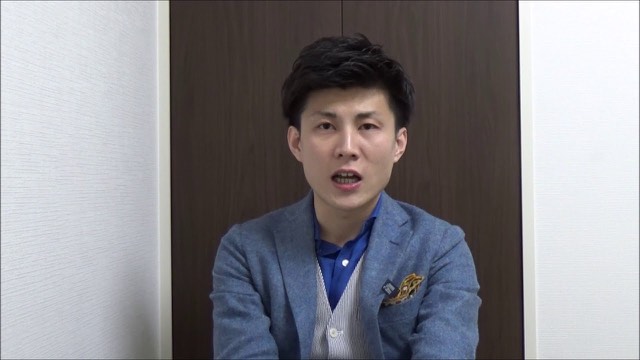 青いブレザーにポケットチーフの本人(木目扉背景)(動画「パチスロ、スロットで勝つスロプロ ハイエナをしてくだ」より)