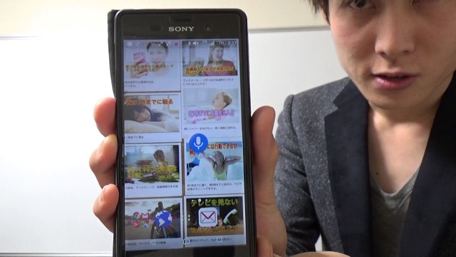 スマホの記事一覧画面を掲げて見せる本人（動画「成功できる人は行動し続ける」より）