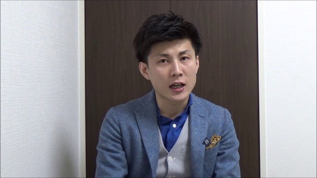 青いブレザーにポケットチーフの本人(木目扉背景)(動画「パチスロ、スロットで勝つスロプロ起業家 期待値が全て」より)