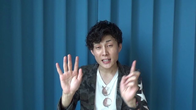 青いカーテン背景で両手を広げる本人(動画「この案件どう思いますか? の見極め方」より)