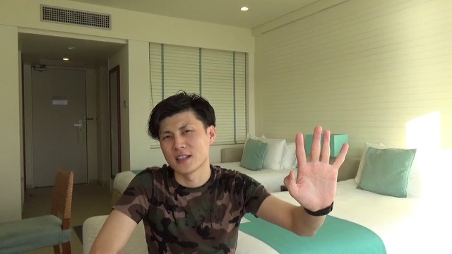 迷彩Tシャツで片手を挙げる本人(ホテル室内)(動画「【パチスロ】勝てない人には3つの理由があります」より)