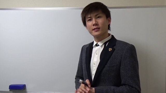 スーツにネクタイ姿でホワイトボード前に立つ本人(なぜ多くの人はパチンコ)(動画「パチスロで負けて金欠=ギャンブル依存症」より)