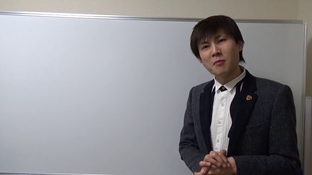 スーツにネクタイ姿でホワイトボード前に立つ本人(動画「借金返済は頑張る理由にならない【パチスロ】…」より)