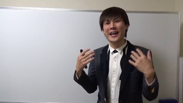 スーツにネクタイ姿で身振りする本人(ホワイトボード前)(動画「パチンコ屋にいるとマインドが腐る【パチスロ】…」より)