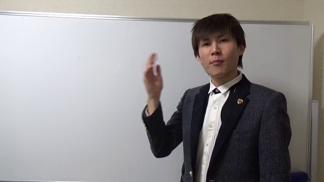 グレースーツでホワイトボード前に立ち片手を挙げる本人(動画「悩めるスロッターへ【パチスロ】【スロット】」より)