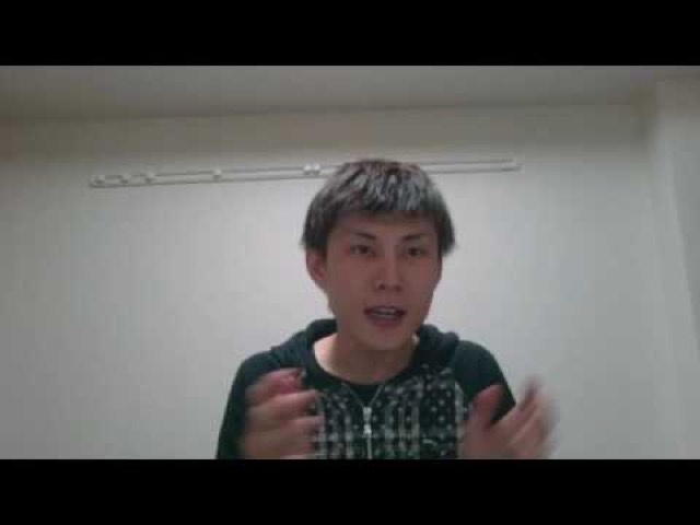 柄物の服を着て無地の壁の前で手を動かしながら話す本人(動画「高設定狙いをする時に意識してること【パチスロ】…」より)