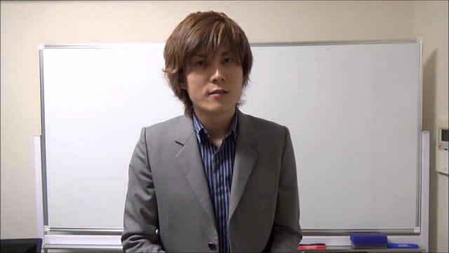 縞シャツにグレースーツでホワイトボード前に立つ本人(動画「自己流は良くない」より)