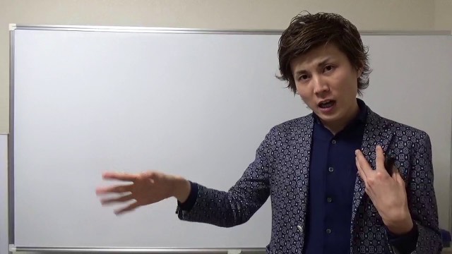 柄物ジャケットでホワイトボード前に立ち片手を伸ばす本人(動画「金持ちになった人がしてた事【パチスロ】【スロット】」より)