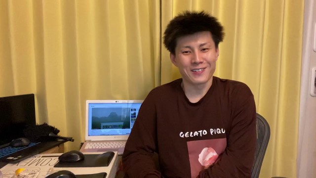 黄色いカーテンを背景に笑顔で腕組みする茶色いパーカー姿の本人