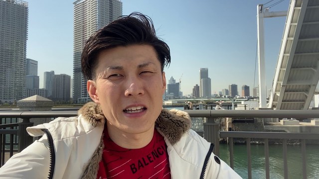 東京の高層ビル群と橋を背景に赤いバレンシアガの服で話す本人(動画「東京で外出自粛が始まりました」より)