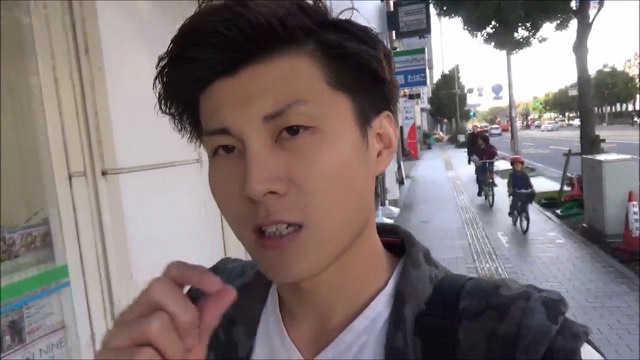 自転車の通行人がいる歩道を背景に話す本人のアップ(動画「ムカつかなくなる方法【ジャグラー】【スロプロ】」より)