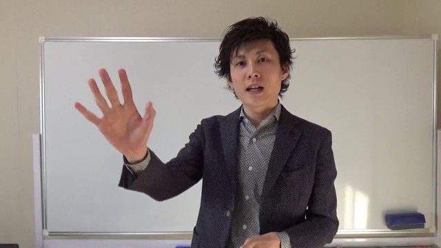 無地の壁を背景に両手で目元を触りながら話す本人(動画「何年経過しても成長しない人【パチスロ】【スロット】」より)