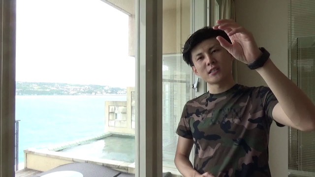 海の見える窓際で迷彩Tシャツを着て話す本人(動画「【答え】パチスロや投資で勝てない理由」より)