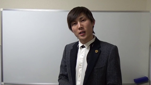 ホワイトボード前でリボンタイ姿で正面を見て話す本人(動画「勝ちたいなら足を使え【パチスロ】【スロット】」より)