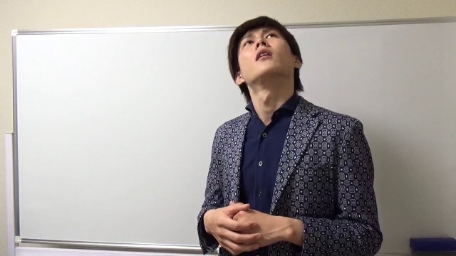 ホワイトボード前で片手を挙げ天井を見上げる柄物ジャケットの本人(動画「グループ打ち=誰でも勝てる それは間違い【パチスロ】」より)