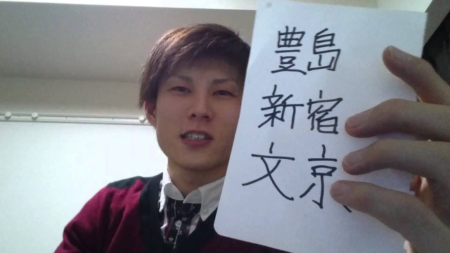 「豊島 新宿 文京」と書いた紙を顔の横に掲げる本人(動画「12月稼働地域抽選」より)