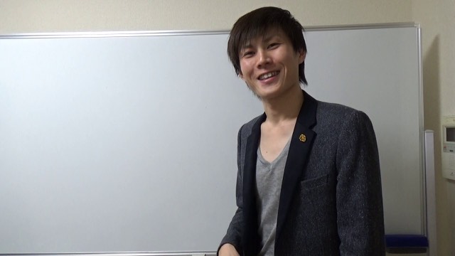 ホワイトボード前で笑顔を見せる金バッジ付きジャケットの本人(動画「初心者向け パチンコ屋には勝てる日がある【パチスロ】」より)